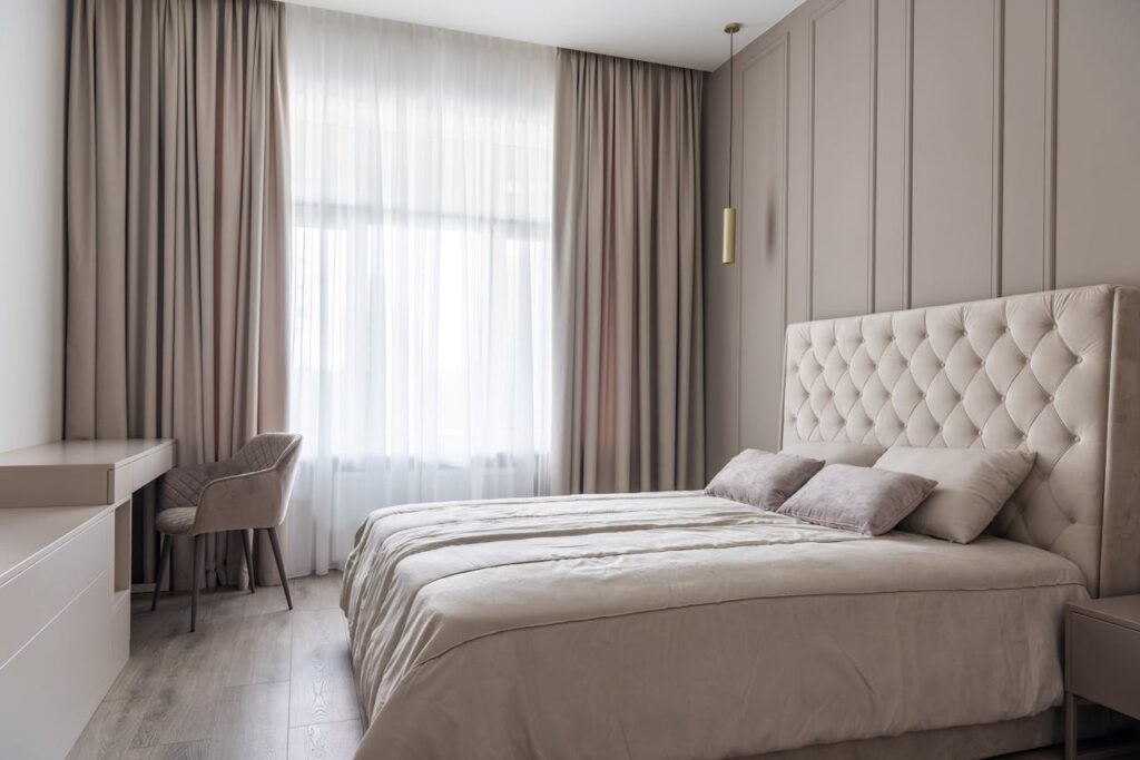 Interior of modern light bedroom with pillows on bed near table with chair next to windows with curtains
