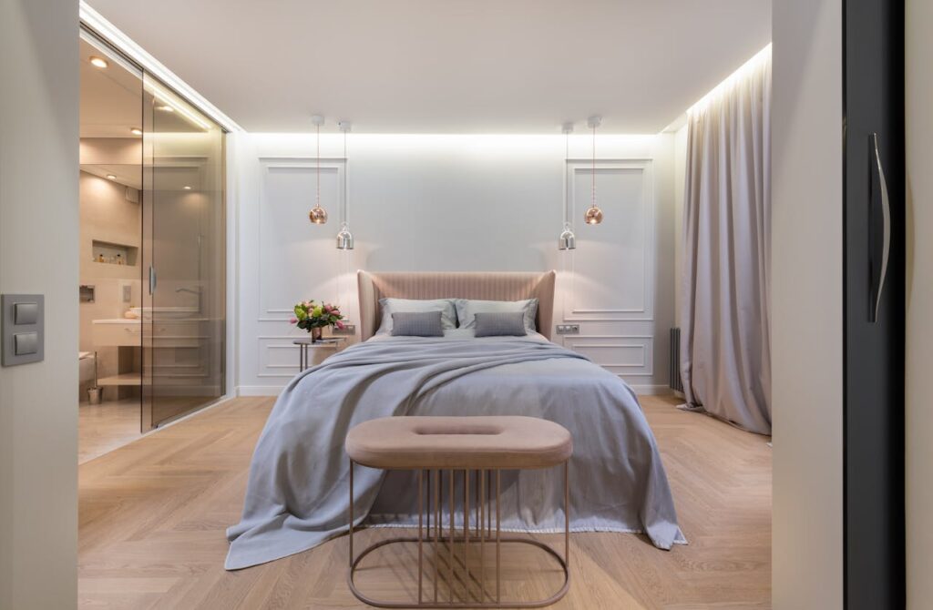 Interior of modern light home with bed with pillows and blanket next to table under lamps near pouf and glass door to washroom